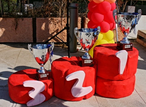 Immagine con rosso, coppe, numeri, podio, tre, palloncini, classifica, bianco