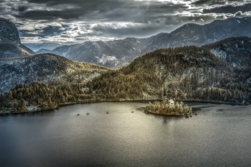 Immagine con lago, neve, nuvole, alberi, isola, acqua, cielo, montagne, inverno, chiesa, boschi, grigio, colline, verde, campanile, riflessi, panorama, monti
