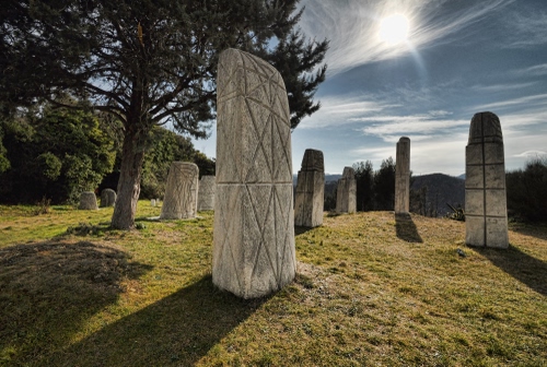 Immagine con cielo, albero, prato, sole, ombre, pietre, erba, stele, pietra, sculture, controluce, monoliti, alberi