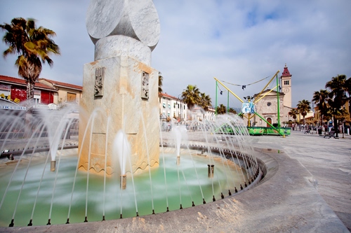 Immagine con fontana, piazza, acqua, chiesa, alberi, zampilli, cielo, nuvole, marmo, palma, palme, getti, spruzzo, spruzzi, pietra, albero, campanile