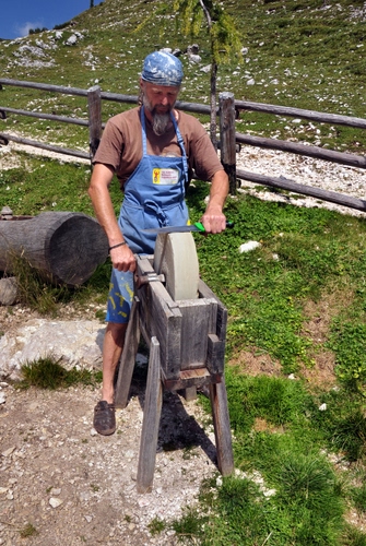 Immagine con arrotino, artigiano, mola, staccionata, uomo, legno, coltello, erba, verde, lavoro, ruota, mani, montagna, barba, tronco