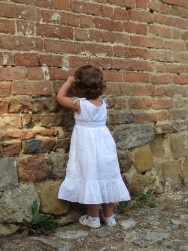 Immagine con muro, bambina, bianco, vestito, nascondino, mattoni, giocare, gioco