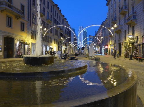 Immagine con fontana, acqua, strada, riflessi, luci, archi, zampillo, case, lampioni, luminarie, via, finestre, palazzi, notte, persone, zampilli, grattacielo, piazza