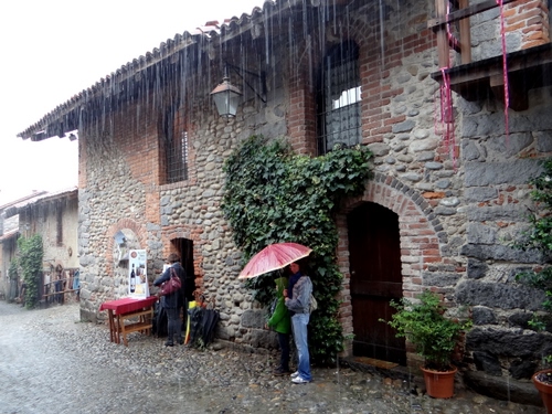 Immagine con pioggia, ombrello, finestre, casa, lampione, vaso, edera, rosso, verde, porta, persone, porte, tavolino, donna, rampicante, maltempo, mattoni, bambino, riparo, piante