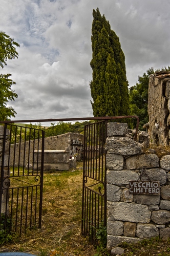 Immagine con cancello, cimitero, nuvole, pietre, cipresso, alberi, tombe, cartello, cipressi, albero, cielo