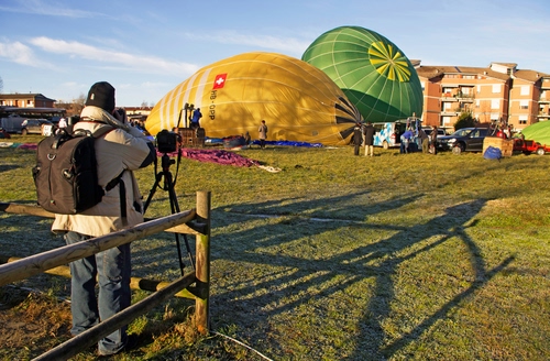 Immagine con mongolfiere, zaino, fotografo, cielo, prato, staccionata, ombra, verde, ombre, giallo, nuvole, fotocamera, uomo, cavalletto, legno, palazzi, case
