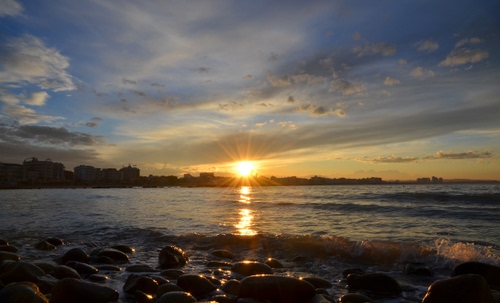 Immagine con tramonto, sole, nuvole, mare, cielo, sassi, alba, riflesso, raggi, onde, acqua, scogli, orizzonte, pietre, città, giallo, spiaggia