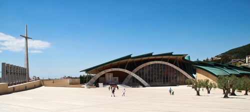 Immagine con croce, alberi, piazza, chiesa, archi, cielo, persone, azzurro, architettura, moderna, tetti, bianco, nuvola, santuario, piazzale