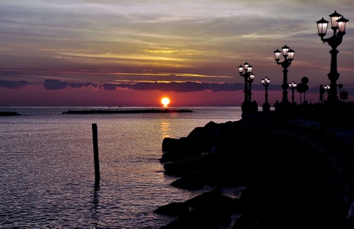 Immagine con mare, lampioni, tramonto, sole, scogli, nuvole