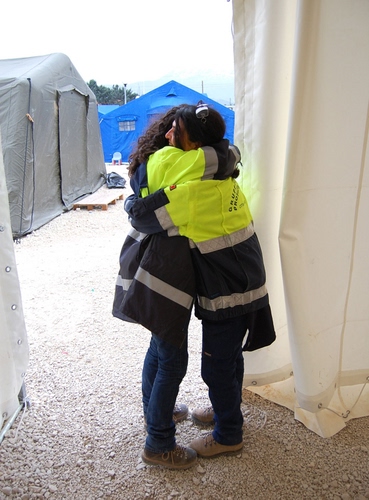 Immagine con abbraccio, tende, giallo, donne, bianco, blu, due, azzurro, grigio, divisa, giacche, occhiali, emergenza, capelli, solidarietà, soccorso, campo, catarifrangenti, sassi