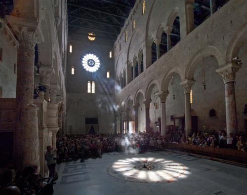 Immagine con rosone, colonne, chiesa, raggi, luce, archi, folla, persone, navata, gente, fedeli, ombra, bari, capitelli, sole, riflesso, finestre, ombre