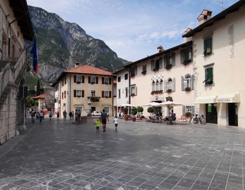 Immagine con finestre, case, montagna, piazza, persone, ombrelloni, bandiera, palazzi, scala, fontana, passanti, fiori, strada, città, persiane, cielo, paese, montagne, tende, porte, nuvole, comignoli
