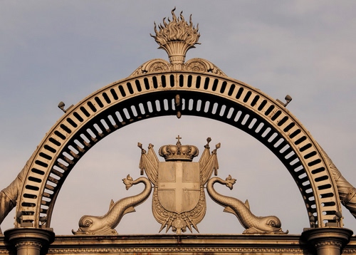 Immagine con stemma, corona, arco, croce, bandiere, pesci, fiamma, fiaccola, delfini, oro, scudo, metallo, cielo, ombra