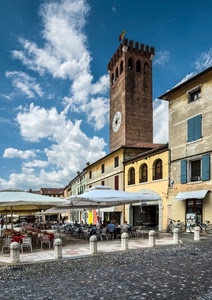 torre, piazza, ombrelloni, orologio, tavolini, sedie, biciclette, nuvole, bandiera, merli, bar, cielo, finestre, palazzi, persone, paracarri, case